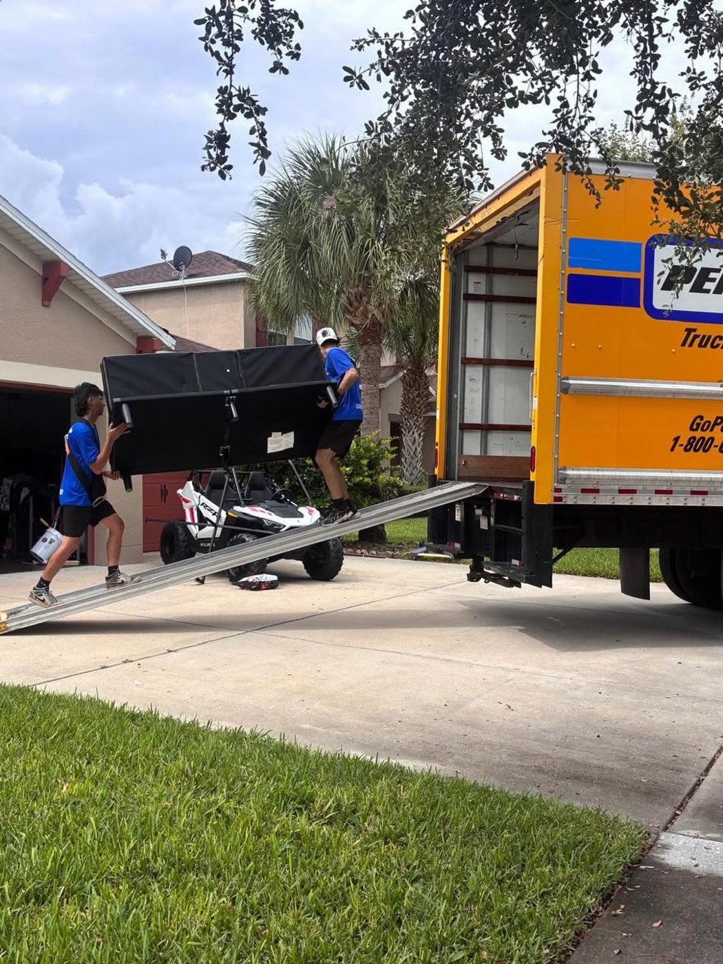 Two crew members carrying a large wrapped item down a truck ramp outside a Florida home
